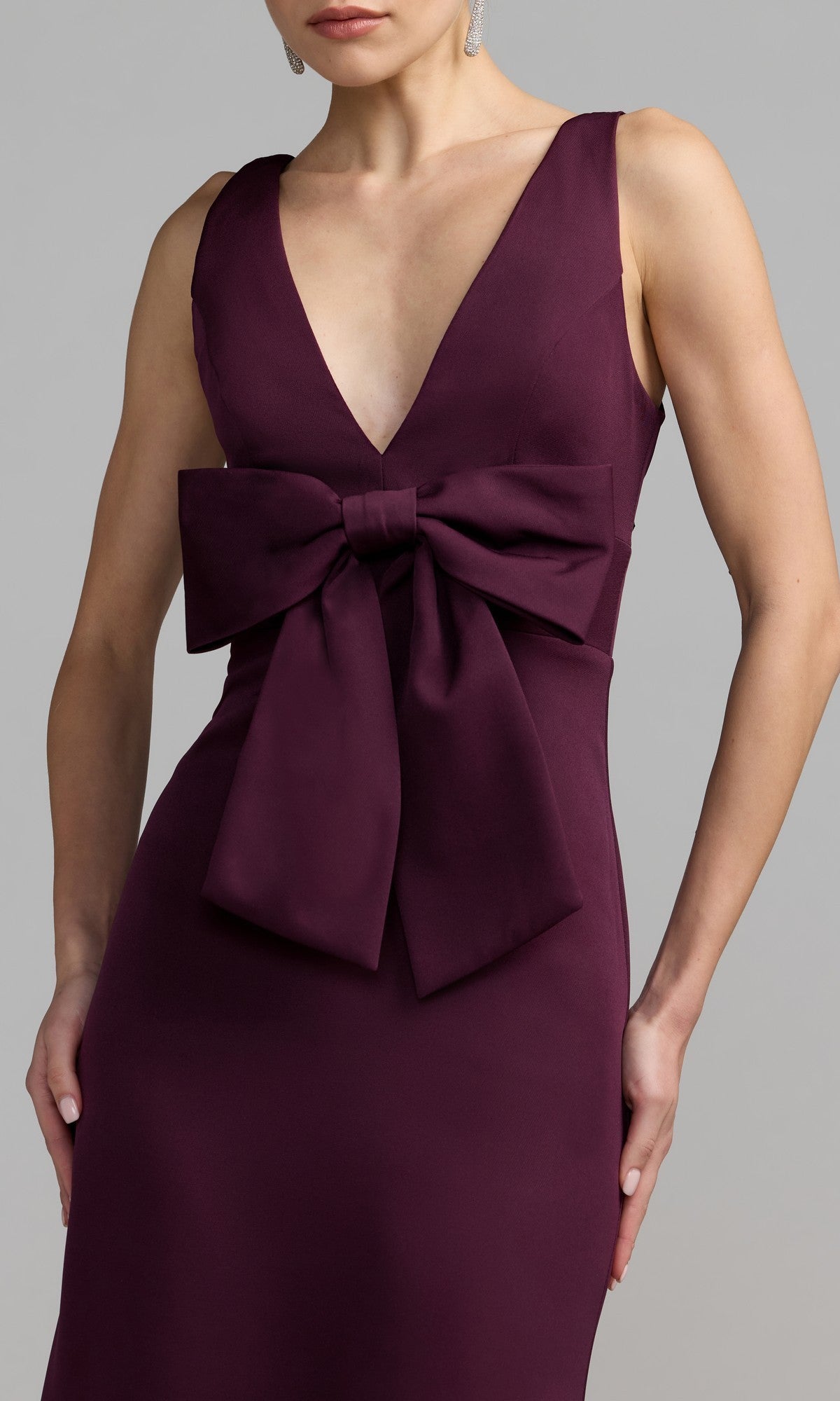A woman poses with one hand on her hip in a sleeveless, deep v-neck Bow-Front Green Formal Dress (Mac Duggal 50087), featuring a large waist bow, against a plain background.